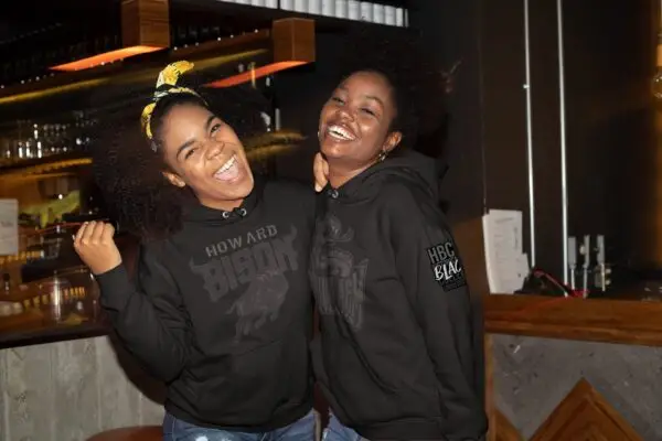 Two young women laughing and embracing wearing blackout Howard University and Florida A&M University Embroidered Patch fleece Hoodies